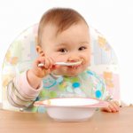 Пятимесячный малыш, сидя в детском стульчике, кушает из тарелки ложечкой